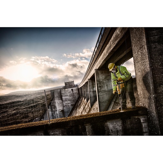 A construction worker wearing a safety helmet and high-visibility jacket uses a DEWALT power tool to drill into concrete at an industrial site with large concrete structures, under a cloudy sky at sunset.