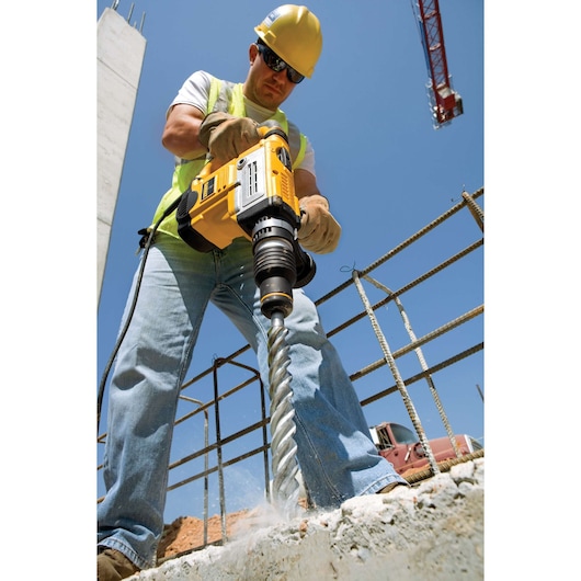 A construction worker wearing safety equipment uses a DEWALT rotary hammer drill on a concrete surface at an outdoor worksite. Metal rebar and construction materials are visible in the background under a clear sky.