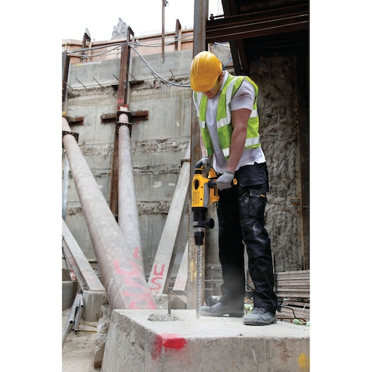 A construction worker wearing a yellow hard hat and safety vest is using a DEWALT rotary hammer drill with the DT9440 bit to drill into a concrete block at a building site.