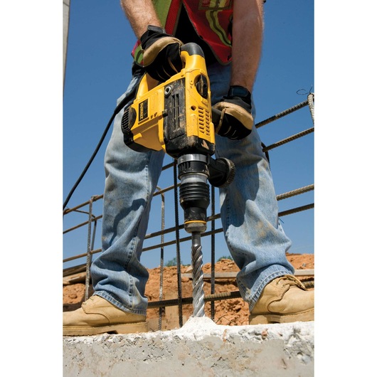 A person using a DEWALT rotary hammer drill with a DT9442 drill bit to drill into concrete at a construction site. The person is wearing gloves, work boots, jeans, and a safety vest.
