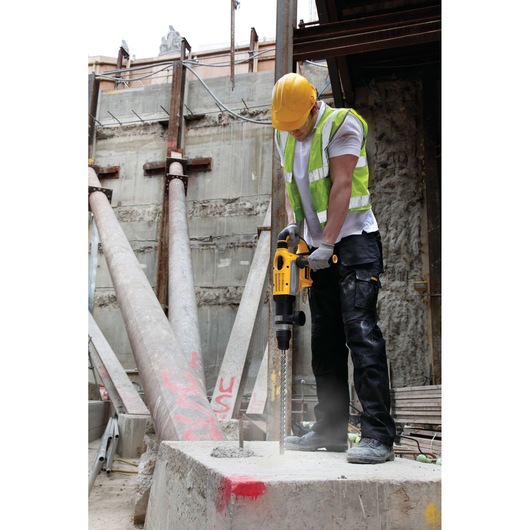 A construction worker wearing a yellow hard hat and reflective vest is using a DEWALT drill on a concrete block at a construction site. Large metal beams and unfinished walls are visible in the background.