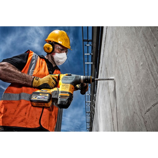 A construction worker wearing a yellow safety helmet, orange high-visibility vest, and gloves uses a DEWALT cordless drill with a drill bit to bore into a concrete wall during daytime.
