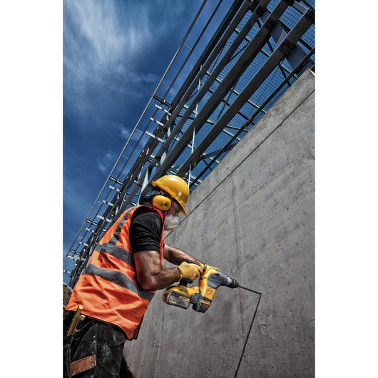 A construction worker wearing a yellow hard hat, ear protection, gloves, and an orange reflective vest is using a DEWALT cordless drill to work on a concrete wall outdoors under a blue sky.