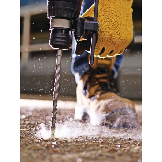 Close-up of a person wearing a yellow work glove operating a drill with a masonry bit, drilling into a concrete surface. Dust and debris are visible around the drill bit. The person is wearing protective work boots.