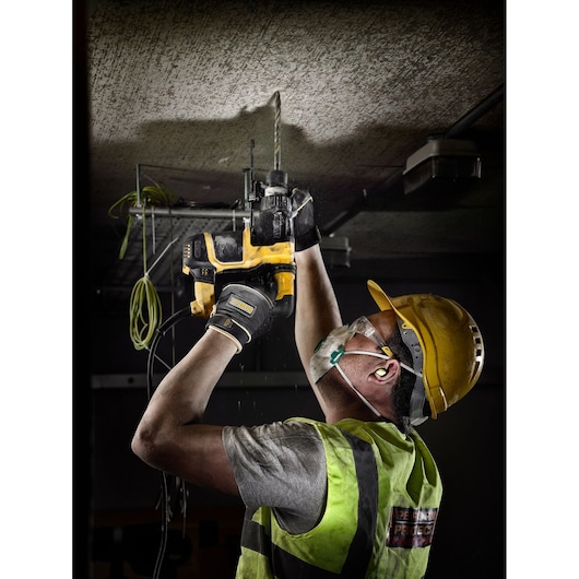 A construction worker wearing a safety helmet, protective vest, gloves, earplugs, and a face mask is using a DEWALT rotary hammer drill (model DT9510) to drill into a concrete ceiling indoors.