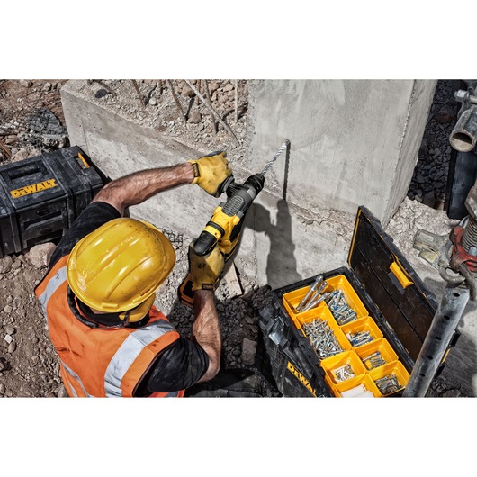 Worker drills concrete wall with DEWALT tool. Tool case with accessories is open beside him.