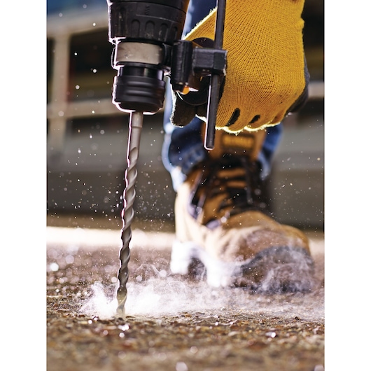 Close-up of a person wearing yellow work gloves and work boots using a drill with a masonry bit to drill into a concrete surface, with dust particles visible around the area.