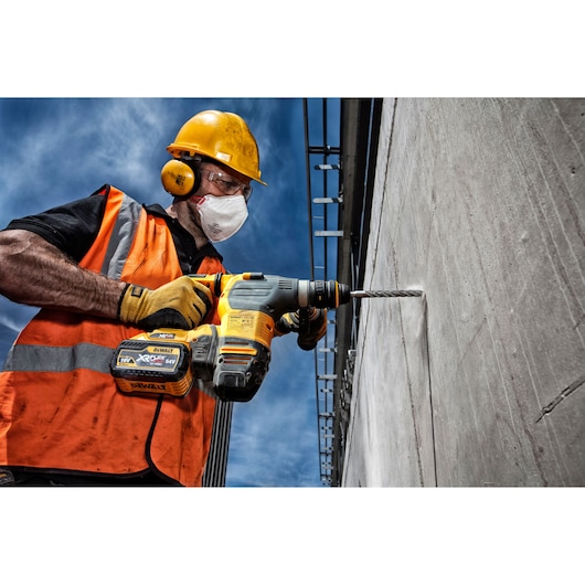A construction worker wearing an orange safety vest, yellow helmet, and gloves is using a DEWALT cordless hammer drill to drill into a concrete wall outdoors under a blue sky.