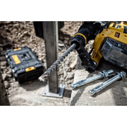 A close-up of a DEWALT rotary hammer drill with a masonry drill bit attached, placed on a concrete surface next to three anchor bolts. In the background, a black and yellow DEWALT tool case is visible on the ground at a construction site.
