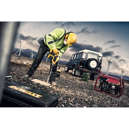 A construction worker wearing safety gear uses a DEWALT power drill to work on rocky ground outdoors. A black utility vehicle, a portable generator, and wind turbines are visible in the background, suggesting a remote or industrial location.