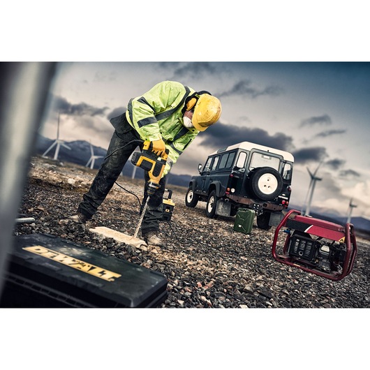 A worker wearing a yellow hard hat and safety jacket uses a DEWALT power drill on rocky ground, with a toolbox labeled DEWALT in the foreground, a utility vehicle, a generator, and wind turbines visible in the background.