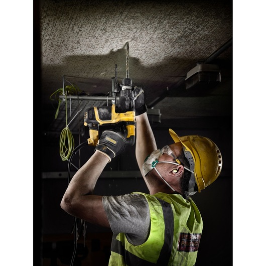 A construction worker wearing a yellow safety helmet, earplugs, a dust mask, gloves, and a high-visibility vest uses a DEWALT power drill with the DT9700 accessory while drilling into a concrete ceiling.