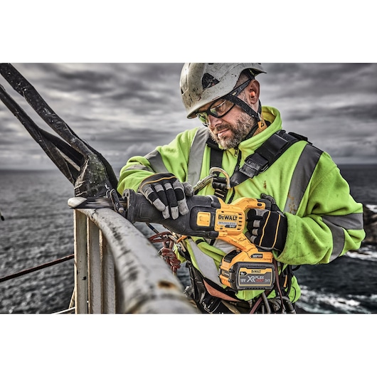 A worker in a high-visibility jacket and safety harness uses a DEWALT cordless reciprocating saw (DT99553) to cut a metal railing outdoors near the sea.