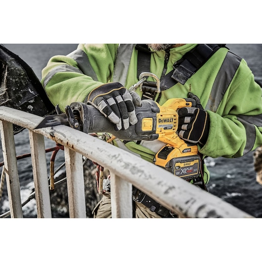 A worker wearing gloves and a green safety jacket uses a DEWALT cordless reciprocating saw to cut through a metal railing outdoors.
