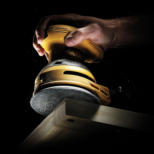 A close-up image of a DEWALT orbital sander being used to sand the edge of a wooden plank, held by a person's hand against a dark background.