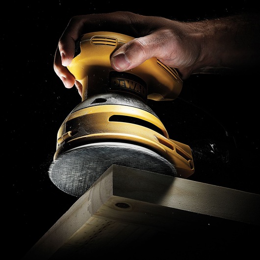 A close-up view of a hand using a DEWALT electric sander on the edge of a piece of wood, with dust visible around the sanding surface.