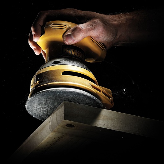 Hand using a DEWALT sander to smooth a wooden plank edge in a dark setting.