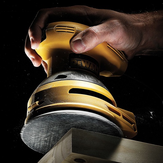 A close-up view of a yellow DEWALT orbital sander being used to sand the edge of a wooden piece. The sanding disc is in motion and dust is visible, with a hand holding the tool.