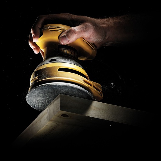 A close-up of a hand holding a yellow DEWALT orbital sander, smoothing the edge of a wooden plank against a black background.