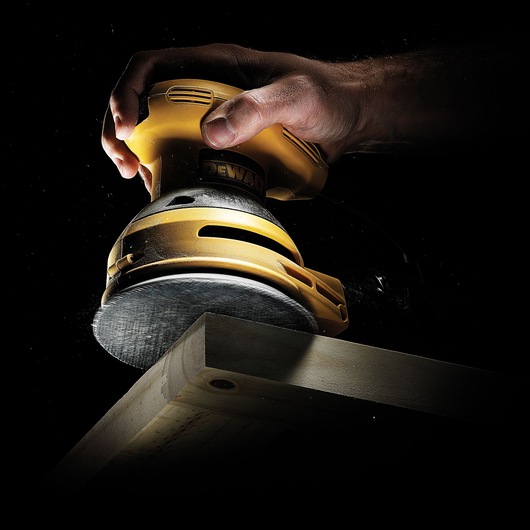 A close-up view of a hand holding a yellow DEWALT power sander, smoothing the edge of a wooden plank against a dark background.