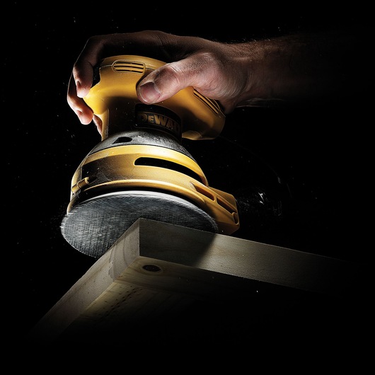 A hand holding a yellow DEWALT electric sander, smoothing the surface of a wooden plank against a dark background.