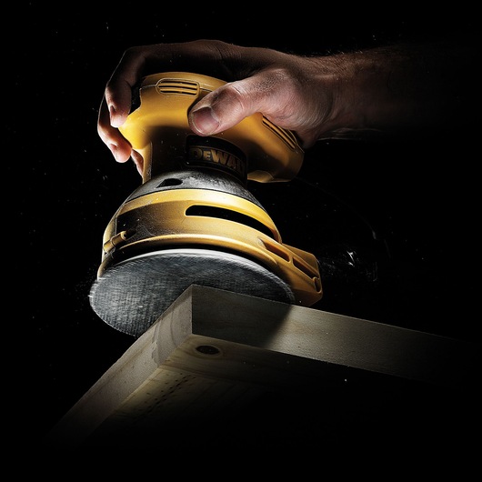 A hand using a yellow DEWALT electric sander to smooth a wooden edge, with dust particles visible against a dark background.