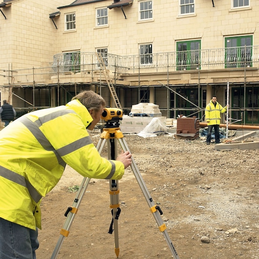 Two workers wearing yellow reflective jackets are using a DEWALT DW096PK automatic level on a tripod at a construction site. One worker is adjusting the device while the other is holding a leveling rod. The site is in front of a building under construction with scaffolding and building materials visible.