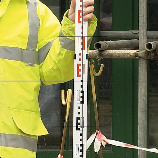 A close-up view of a person wearing a high-visibility yellow jacket, holding a leveling staff for surveying, with construction materials visible in the background.
