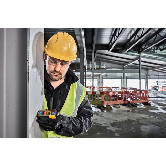 A construction worker wearing a yellow hard hat, safety vest, and gloves is holding a DEWALT DW099S laser distance measurer against a metal beam in an industrial building under construction.