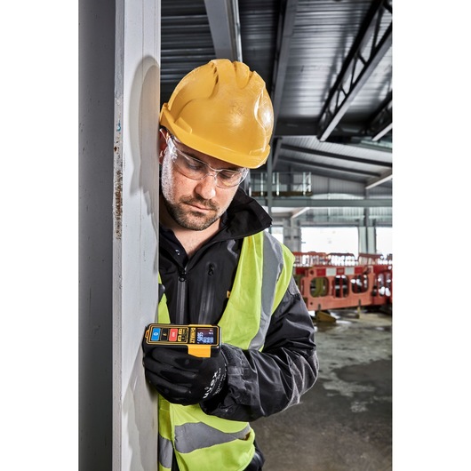 A person wearing a yellow hard hat, safety vest, and black gloves is using a DEWALT DW099S laser distance measurer against a vertical surface in an industrial indoor construction site.