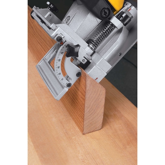 Close-up of a DEWALT biscuit joiner tool in use, with its adjustable fence set at an angle, positioned on a piece of wood for precise joinery.