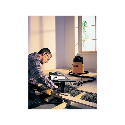 A person using a DEWALT DW711 saw to cut wooden planks during construction work, with a DEWALT yellow vacuum connected for dust extraction, indoors near a window.