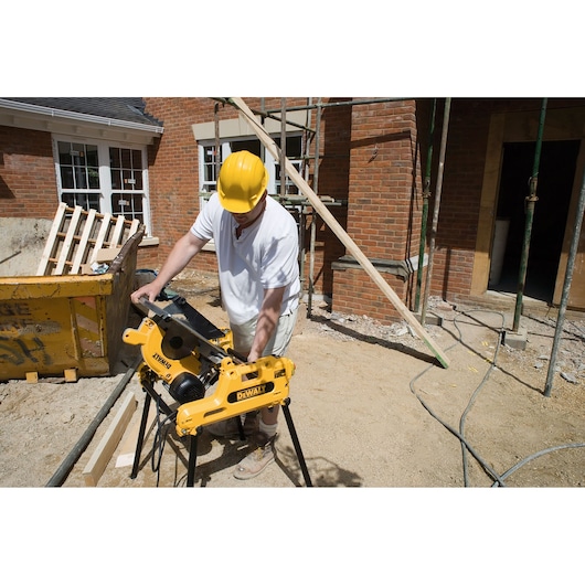 A construction worker wearing a yellow hard hat operates a DEWALT DW743N saw on a building site with brick walls and scaffolding in the background.