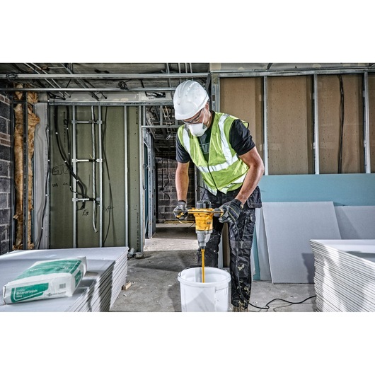 A construction worker wearing a safety helmet and high-visibility vest is using a DEWALT DWD241 mixer to blend materials in a white bucket inside a building under construction. There are stacks of drywall panels and a package of materials nearby.