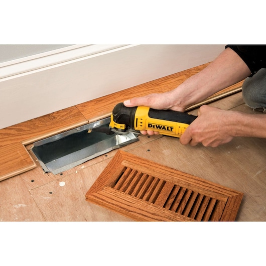 A person using a DEWALT DWE315KT oscillating multi-tool to cut or adjust wooden flooring around a metal vent opening. An unattached wooden vent cover is placed nearby.