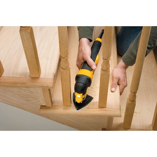 A person using a DEWALT yellow and black multi-tool to sand or cut wood on a staircase, focusing on working between wooden balusters.