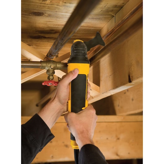 A person uses a yellow DEWALT oscillating multi-tool to cut a metal pipe under a wooden ceiling structure.