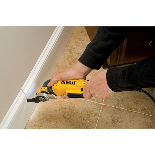A person using a yellow DEWALT oscillating multi-tool to cut a white baseboard along a tiled floor.