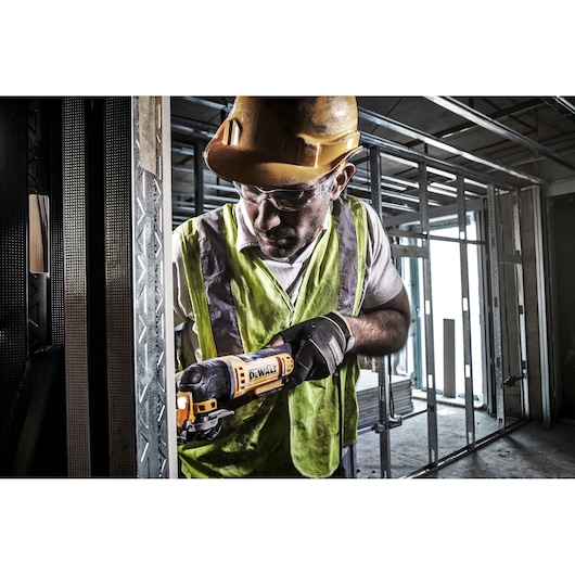 A construction worker wearing a safety vest and hard hat uses a DEWALT DWE315 oscillating tool to cut metal framing inside a building under construction. The worker's face is blurred for privacy.