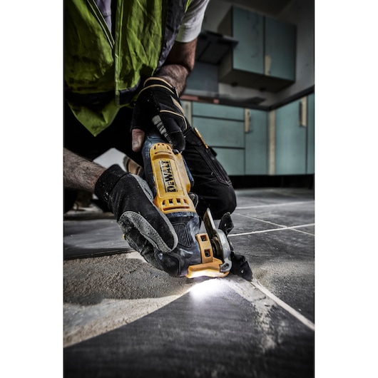 A person wearing work gloves and a safety vest uses a DEWALT DWE315 oscillating multi-tool to cut grout between floor tiles in a kitchen setting, with a bright light illuminating the cutting area.