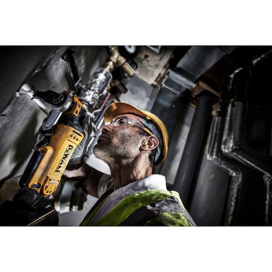 A worker in safety gear uses a DEWALT DWE315 power tool to cut into a wall in an industrial setting. The worker's face is blurred for privacy.