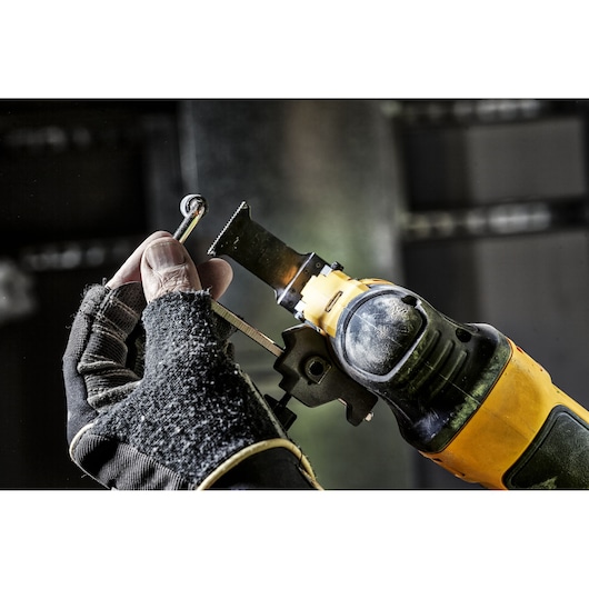 A hand wearing a work glove is holding a metal object near the blade of a yellow and black DEWALT oscillating multi-tool.