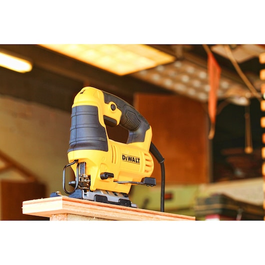 A yellow and black DEWALT electric jigsaw is positioned on the edge of a piece of wood in a workshop setting.