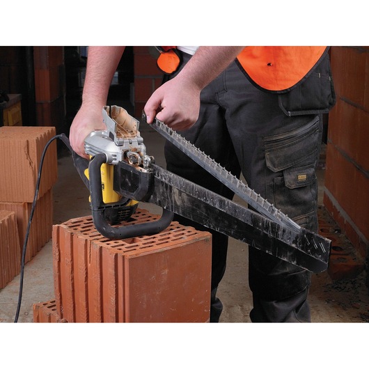 A person using a DEWALT electric masonry saw to cut a brick block. The saw is powered and has a long blade designed for cutting brick or masonry materials, and the person is wearing a construction vest.