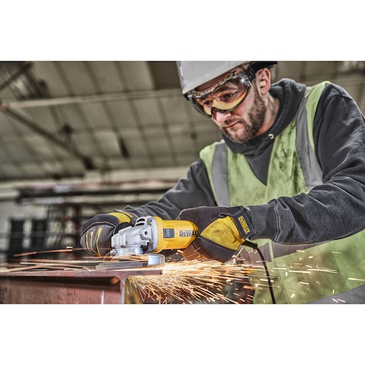 A person wearing safety gear is using a yellow DEWALT angle grinder to cut or grind metal, producing sparks, in an industrial setting.