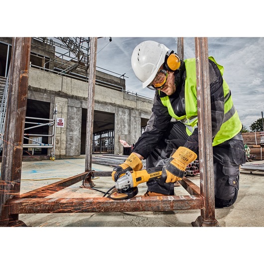 A construction worker wearing protective gear, including a helmet, ear protection, gloves, and a high-visibility vest, uses a DEWALT angle grinder (model DWE4357) to grind a rusty metal frame at a construction site, with sparks flying.