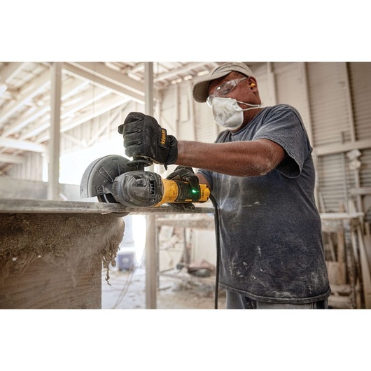 A worker wearing protective gear, including gloves, a mask, goggles, and earplugs, is using a DEWALT angle grinder to cut a piece of material in an industrial workshop setting.