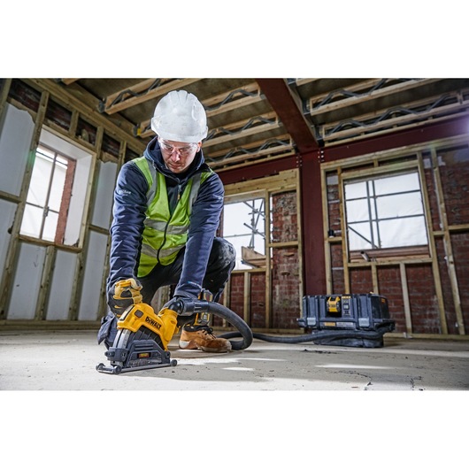 A construction worker wearing a hard hat and safety vest uses a DEWALT power tool (DWE46225) to cut or grind a concrete floor inside a building under construction. A DEWALT equipment case is visible in the background.