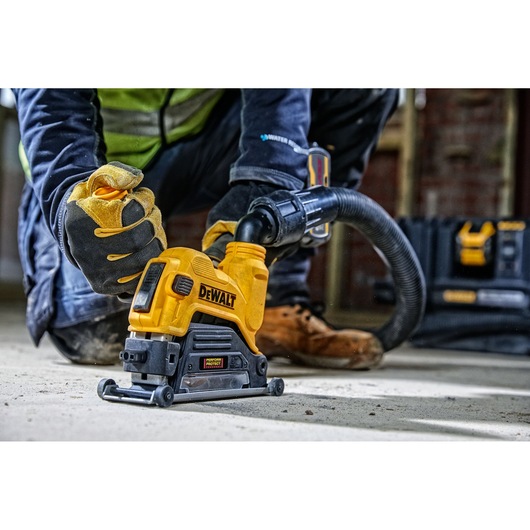 A person wearing safety gloves and work gear using a DEWALT power tool, specifically a surface grinder, connected to a dust extraction hose on a concrete floor at a construction site.
