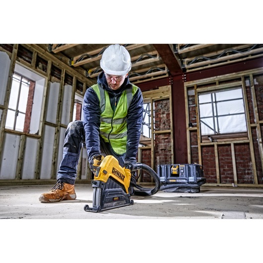 A construction worker wearing a white hard hat, green safety vest, and work boots is using a DEWALT power tool on the floor of an unfinished building with exposed wooden framing and brick walls. The worker's face is blurred. A DEWALT toolbox is visible in the background.